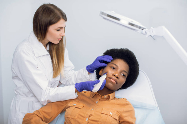 Beauty and face care concept. Female doctor cosmetologist in blue gloves makes ultrasound cleaning procedure of facial skin of young pretty african woman in beauty salon. - 写真・画像