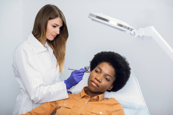 Cosmetology and beauty concept. Close up portrait of pleasant African woman getting facial oxygen anti-aging procedure in beauty salon. European female doctor makes needle free biorevitalization. - Foto, immagini