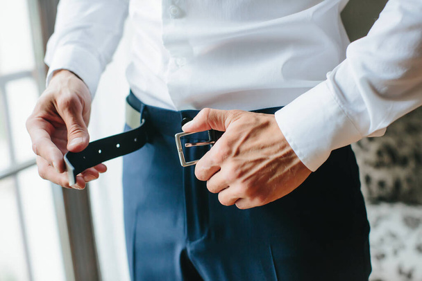 A young man fastens a black belt on his trousers, male hands close-up. Morning of the groom, preparations for the wedding. Selective focus. - Foto, Imagem