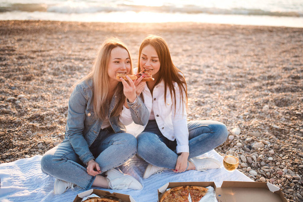 Dwie młode piękne dziewczyny bawiące się pizzą i winem na plaży nad brzegiem morza w świetle słonecznym. Letni sezon wakacyjny. Przyjaźń. Szczęście. Siostry spędzają razem czas na wybrzeżu.. - Zdjęcie, obraz