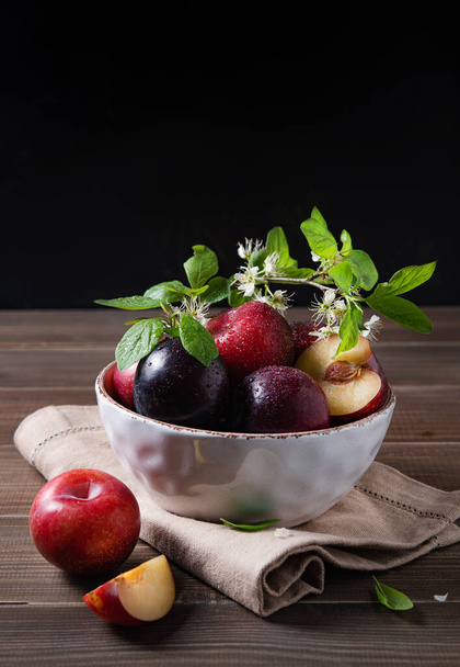 a few juicy red plums in bowl with branch blossom flowers on a napkin on a brown wood background. Front view  and copy space image - Foto, Bild