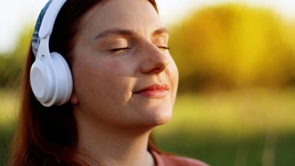 Beautiful woman with closed eyes listening to music using wireless headphones and smartphone in city park - 写真・画像