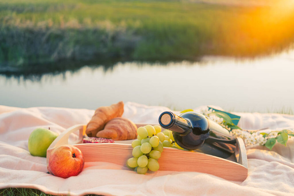 Picknick bij de rivier. Op een licht gekleurde sprei, een houten dienblad met fruit, gebak en een fles wijn. Het licht van de ondergaande zon, een aangename rustige sfeer, heldere kleuren. - Foto, afbeelding