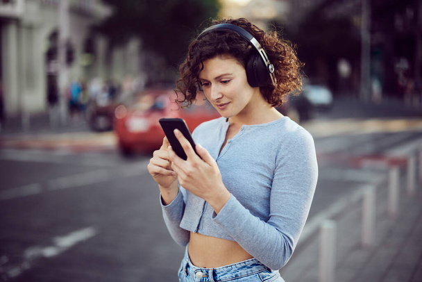 Cute girl with curly hair standing on the street with headphones on and choosing music. - Photo, Image