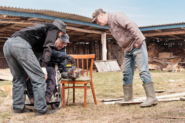 Un homme âgé en fauteuil roulant est en train de réparer une vieille scie électrique dans sa cour. Ses deux amis regardent attentivement. - Photo, image