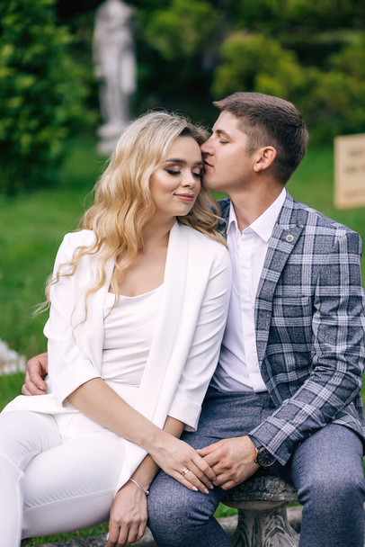 Stylish groom in a gray checkered suit and a cute blonde bride are hugging while sitting on a background of greenery in the park. - Zdjęcie, obraz