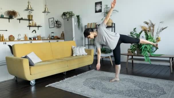 Una giovane donna indiana fa meditazione yoga a casa, fa un esercizio di equilibrio, sta su una gamba in una stanza luminosa, dietro un divano giallo e la cucina, la ragazza è vestita con una t-shirt bianca e gambali neri - Filmati, video