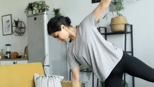 Una joven india hace meditación de yoga en casa, hace un ejercicio de equilibrio, se para en una pierna en una habitación brillante, detrás de un sofá y cocina amarilla, la chica está vestida con una camiseta blanca y leggings negros - Metraje, vídeo