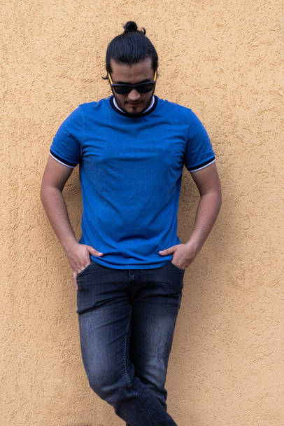 Mladý Latinoameričan se slunečními brýlemi a rukama v kapsách, stojící na hořčičně zbarvené zdi a dívající se dolů. Čerstvý koncept - Fotografie, Obrázek