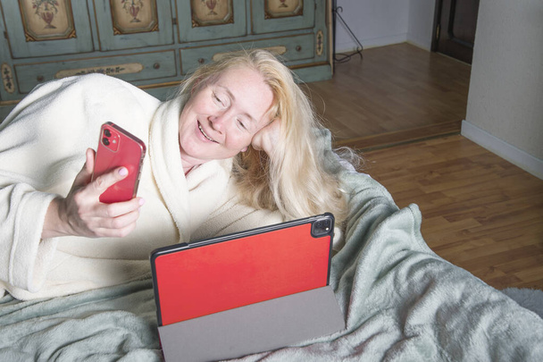 eine lachende blonde Frau mittleren Alters mit langen Haaren im Bademantel liegt auf dem Bett und spricht auf einem Smartphone - Foto, Bild