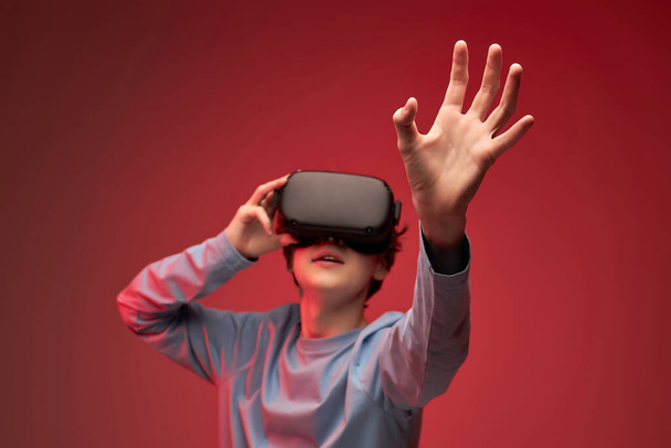 Portrait of young teen boy in VR helmet looks away at copy space, isolated on red - Foto, immagini