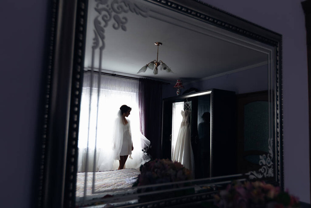 odbicia w lustrze, smukła kobieta w szlafroku z welonem stoi obok łóżka i patrzy na suknię ślubną. - Zdjęcie, obraz