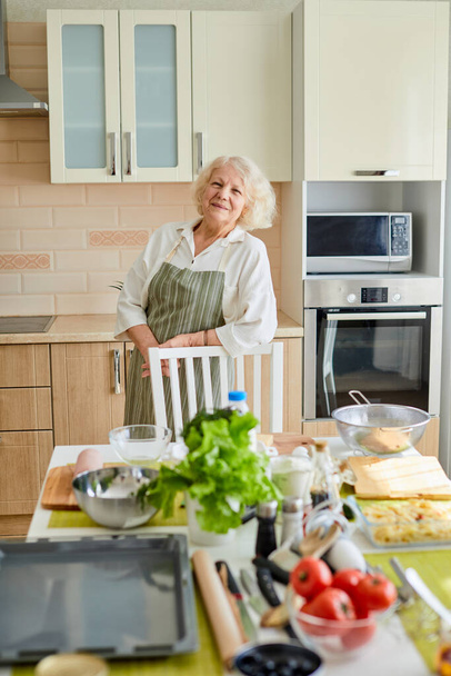Agradable dama de pelo gris en delantal durante la cocina en casa, sonriendo - Foto, imagen