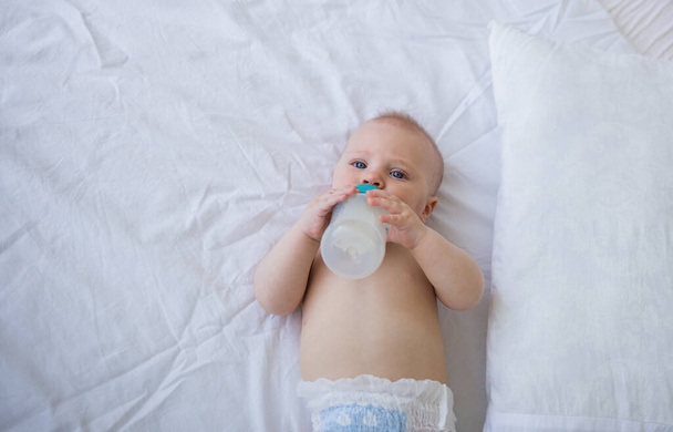 Baby Junge mit blauen Augen in Windel liegt auf dem Bett und hält eine Flasche mit Formel. Künstliche Ernährung - Foto, Bild
