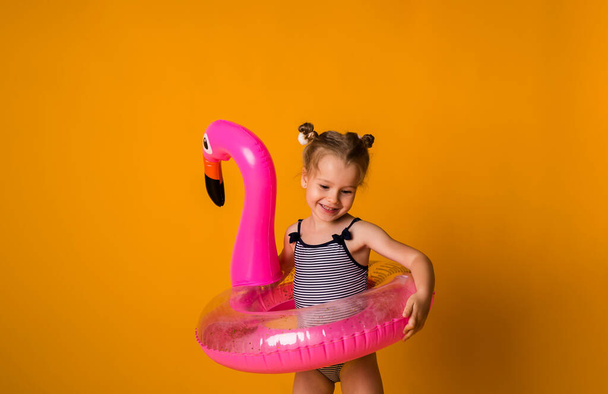 счастливая маленькая блондинка в полосатом купальнике держит розовый надувной круг на желтом фоне с копией пространства - Фото, изображение