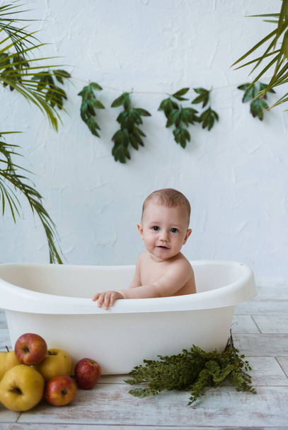 Το αγοράκι λούζεται σε ένα λευκό μπάνιο με μήλα σε λευκό φόντο με φυτά. Θερινό φόντο - Φωτογραφία, εικόνα