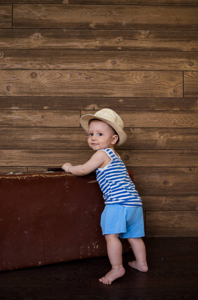 маленький мальчик стоит рядом с ретро чемоданом на коричневом деревянном фоне с местом для текста. Летние каникулы - Фото, изображение
