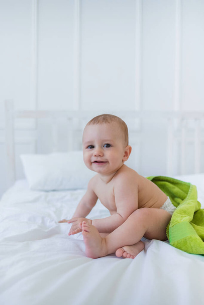 bebé niño con una toalla verde se sienta en una manta de algodón blanco en una cama con espacio para el texto - Foto, imagen