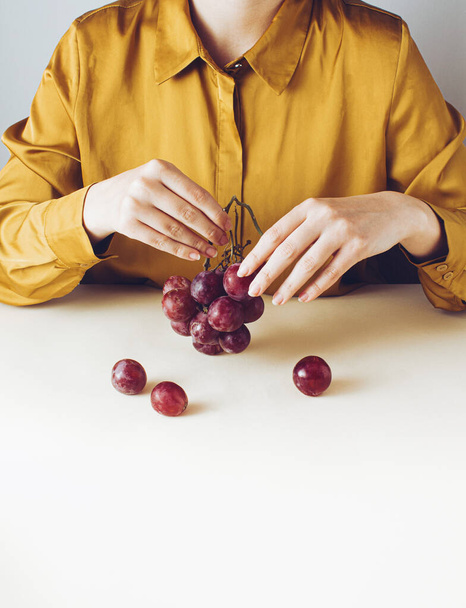 Composizione unica nella vita reale. Donna elegante con uva rossa in mano. Concetto minimale stile retrò. - Foto, immagini
