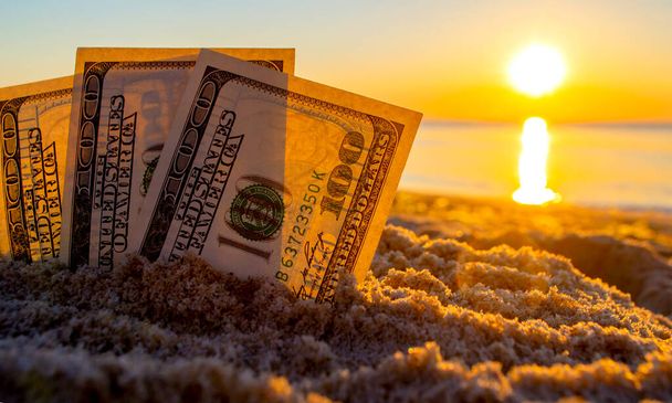 Três notas de dólar são enterradas na areia na praia perto do mar ao pôr-do-sol - Foto, Imagem