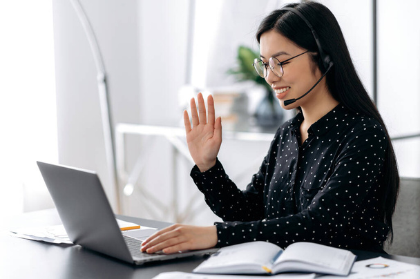 Friendly beautiful smart asian woman wearing glasses and headset, freelancer, support employee or manager sitting at table in office talking to client or employee, waving hand, smiling - Photo, Image