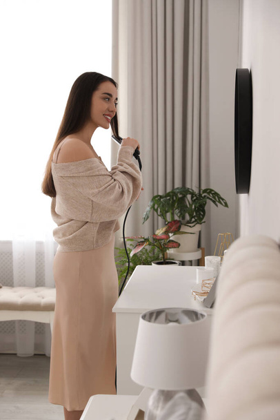 Young woman straightening hair near mirror at home. Morning routine - Foto, afbeelding