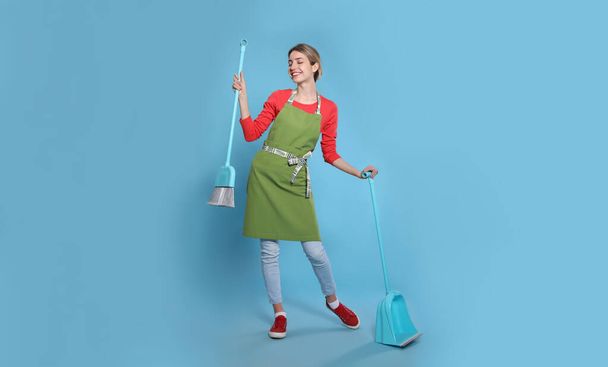 Young housewife with broom and dustpan on light blue background - Фото, изображение