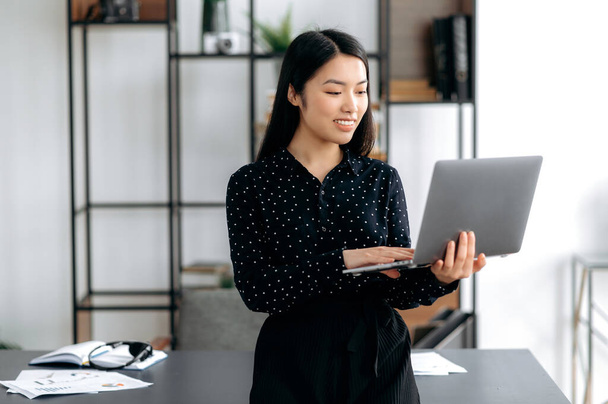 Portrait attractive confident successful smart pretty young asian business lady or manager standing near work desk in office, in formal clothes, holds a laptop in hands, browsing news, smiles - Photo, Image
