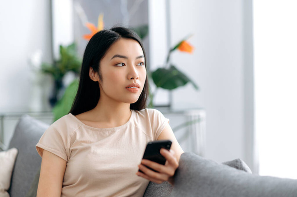 Pensive young asian beautiful woman uses her mobile phone while sitting on the sofa in casual clothes, browses the internet and social networks, texting with friends or family, looks away thoughtfully - Φωτογραφία, εικόνα