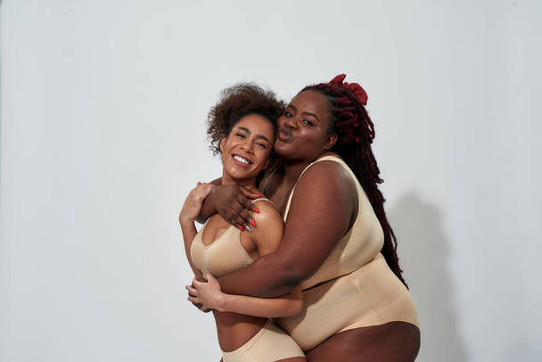 Dvě hravé, zakřivené a štíhlé africké americké ženy ve spodním prádle objímající a zároveň pózují společně pro kameru izolované přes šedé pozadí - Fotografie, Obrázek