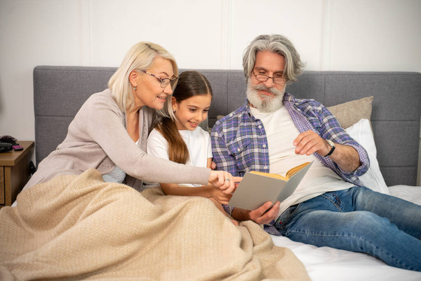 Улыбающиеся старшие бабушка и дедушка читают книгу с маленькой девочкой, лежащей на кровати - Фото, изображение