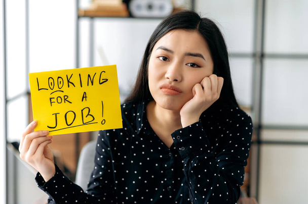 Sad unemployed young clever asian woman, student, sits at a desk with sign with the inscription looking for a job, hopes to get a dream job, asks for support, sadly looks at camera - Photo, Image