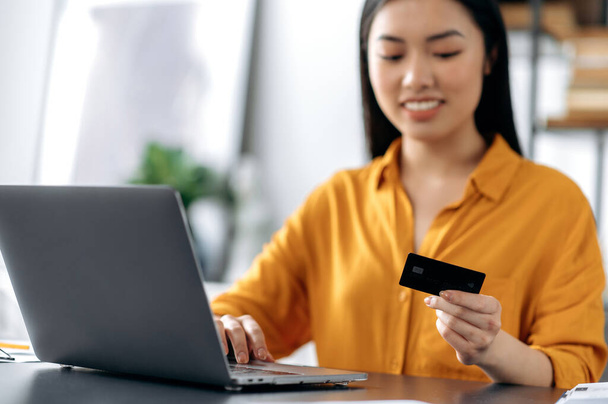 Online shopping. Happy blurred beautiful young asian brunette woman sitting at the table, uses laptop and credit card to pay for online purchases and delivery, enters data, looks at the card, smiles - Photo, Image
