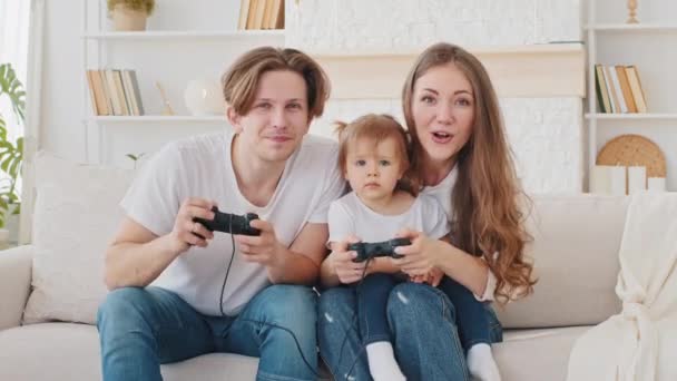 Καυκάσιος οικογένεια παίζει κονσόλα βιντεοπαιχνιδιών στο σπίτι κάθεται στον καναπέ νεαρή μητέρα και το μικρό παιδί κόρη μικρό κορίτσι κερδίσει ηττημένος πατέρας ανταγωνισμού αστείο αναψυχή, ζευγάρι με το μωρό απολαμβάνουν videogaming - Πλάνα, βίντεο