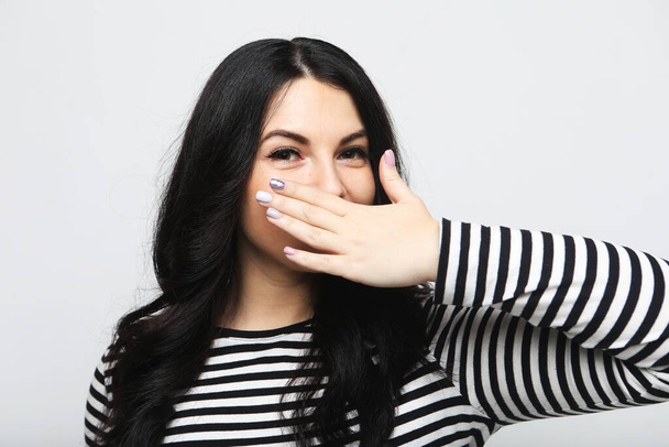 Body language, emotions, and people concept: Young brunette female in a striped sweater smiles and is surprised, covering her mouth with her palm - Foto, Imagen
