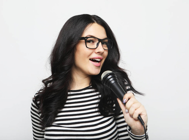 Jovem morena de camisa listrada e óculos segura microfone e se prepara para cantar em karaoke sobre fundo cinza - Foto, Imagem