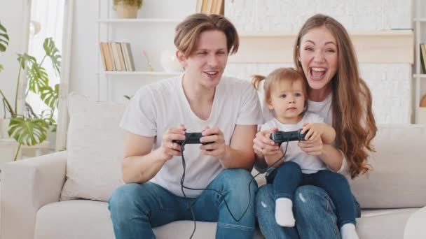 Caucasian young millennial family man father and beautiful mother woman playing video game console at home sitting on couch, mom holding little daughter child girl win cheering in videogaming applaud - Footage, Video