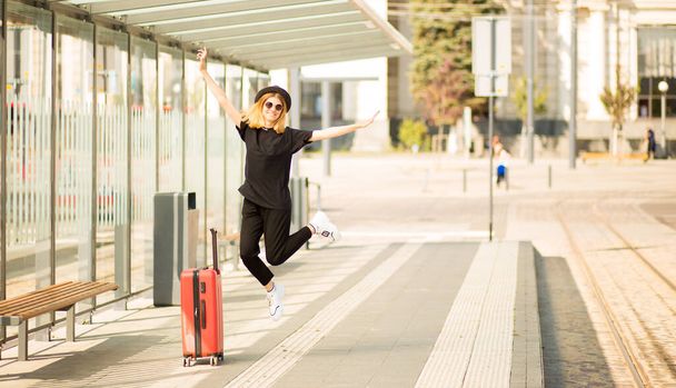 Innoissaan turisti nainen pukeutua täysin musta matkatavarat hyppäämällä raitiovaunuasemalla. Iloinen tyttö matkaaja hypätä matkalaukku. Matkakonsepti Nauti avoimista rajoista turvallinen maa lähtee  - Valokuva, kuva