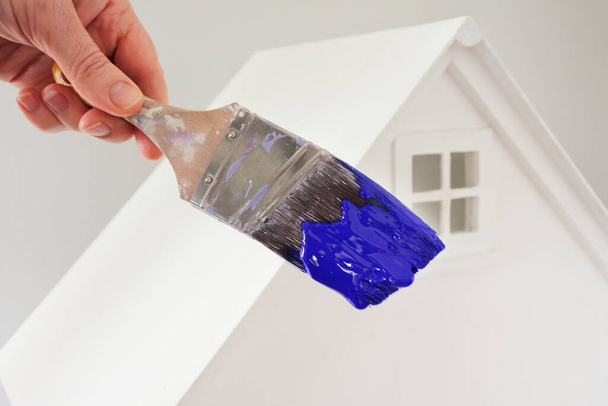 Pessoa segurando um pincel de tinta com cor de tinta azul contra uma casa branca. Renovação, decoração de casa e pintura casa conceito. Não há pessoas. Espaço de cópia - Foto, Imagem