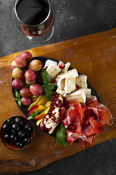  A set of appetizers for wine, jamon, pepperoni, cheese, grapes, peach and olives on a wooden board top view. Snack board and a glass of red wine on dark gray background, vertical photo - Photo, image