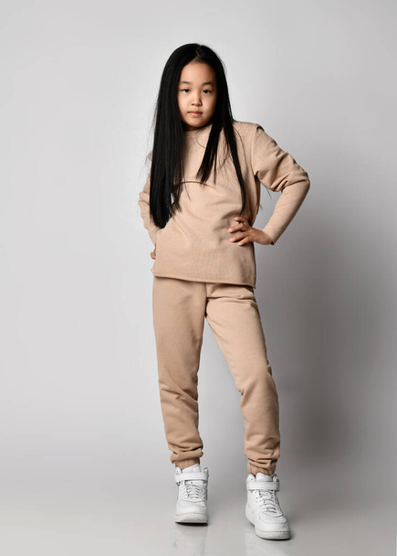 Cool Asian kid girl green pants, hoodie, sweater and sneakers stands holding hand at waist, posing in new sportswear - Foto, Imagem