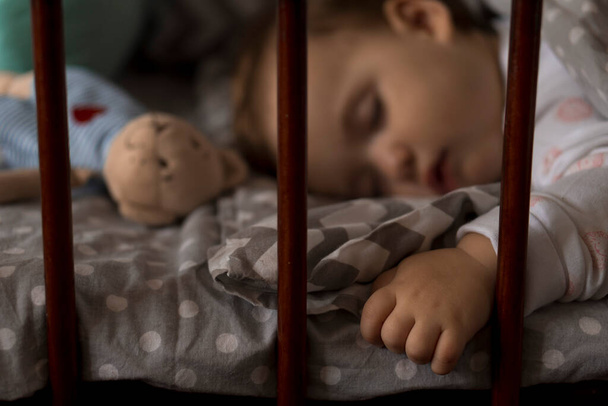 Милая весёлая маленькая пухленькая девочка, сладко спящая в серой кроватке во время обеденного отдыха в белой и розовой пижаме с плюшевым мишкой дома. Детство, отдых, комфорт, медицина, концепция здоровья - Фото, изображение
