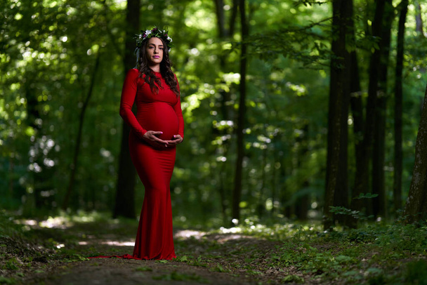 Portrait of a beautiful young hispanic pregnant woman in long red dress in a deciduous forest in the summer - Fotografie, Obrázek