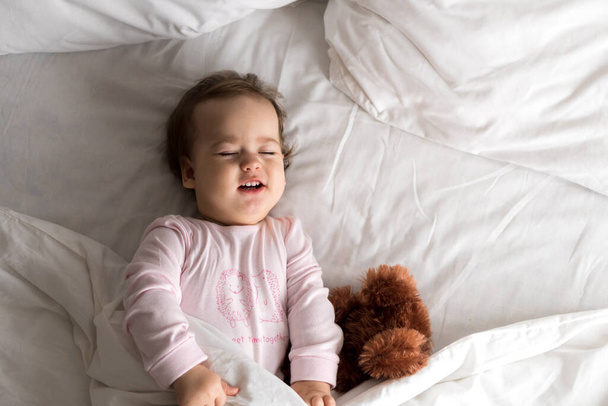 Autentický portrét roztomilé kavkazské malé dítě baculaté holčička nebo chlapec v růžové ospalý po probuzení s medvídkem dívá na kameru v bílé posteli. Péče o děti, Dětství, Rodičovství, koncept životního stylu - Fotografie, Obrázek