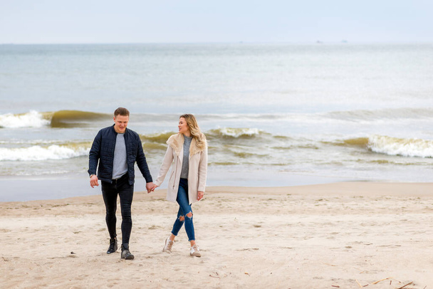 Attractive young couple walking along the shore of a sandy beach, on a spring romantic holiday, outdoors. Travel tourists lifestyle. Couple enjoying loving time together. - Zdjęcie, obraz
