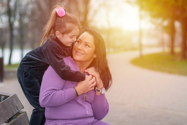 Little cute girl hugging her mother affectionately from behind in a park.Summer spring sunset park background. - Photo, Image
