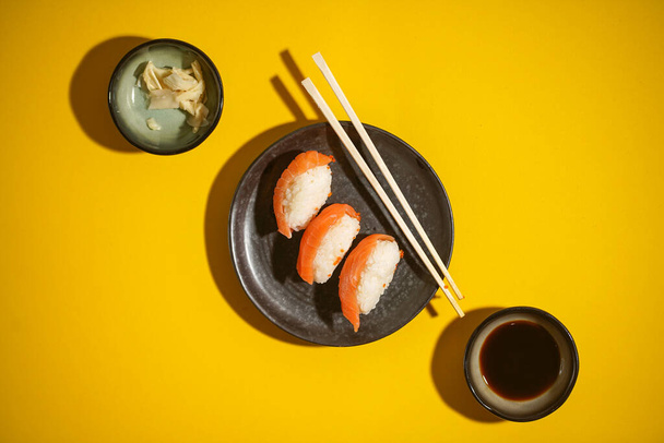 Sushi nigiri z łososiem na czarnym talerzu na żółtym tle, z sosem sojowym i imbirem, widok z góry - Zdjęcie, obraz