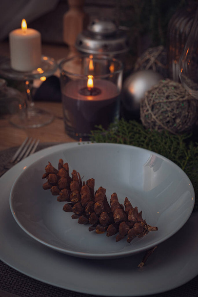 A conifer cone in a bowl on a table with a burning candle in a glass and Christmas decorations - Foto, imagen