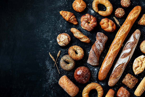 Bakery - various kinds of breadstuff. Bread rolls, baguette, bagel, burger bun and croissant captured from above (top view, flat lay). Black chalkboard background with free copy (text) space. - Фото, изображение