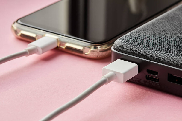 El banco de energía carga el teléfono inteligente usando un cable USB en una superficie rosa. Primer plano, enfoque selectivo - Foto, Imagen
